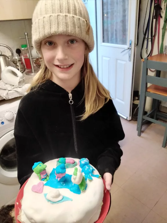 Mina is holding a cake covered in white icing. She is wearing a black cardigan and a winter hat.