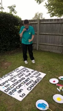 Carly is standing in a garden, looking down at a big sign that reads 'Participation is having opportunities to be part of decisions that are important to you and may lead to changes'.