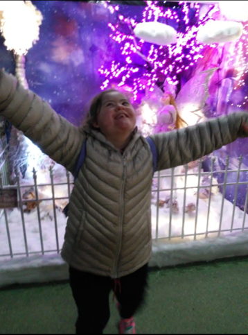 Taylor is at an event. She is looking up at the sky with both arms stretched out. It looks like winter and there are Christmas lights in the background.