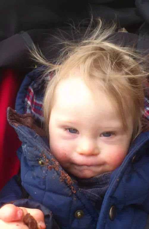 Ollie is photographed in his car seat. He is wearing a blue coat and is eating a snack. His hair is disheveled. 