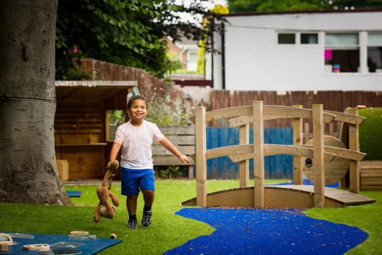 Joseph is walking in a garden, holding a teddy with one hand. He is wearing a pink t-shirt and blue shorts.
