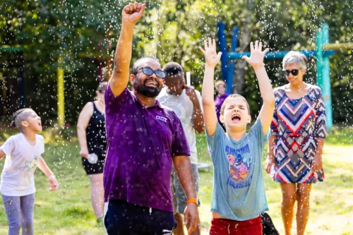 A staff member and child are standing outside with their arms up in the air and water drops falling on them
