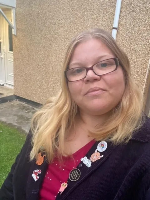 Allie is taking a selfie. She is wearing a black jackets with pins on it.