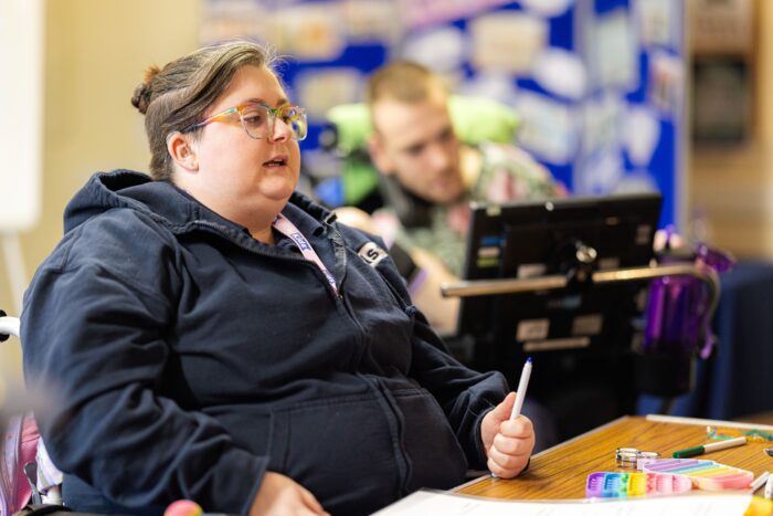 A person is sitting around a wooden table, a pen in their hand. They are looking at someone off camera, presumably talking to them
